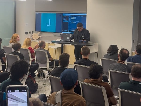 A photograph capturing an indoor technology presentation or lecture, showing a male presenter standing next to a table with a laptop, addressing an audience seated in rows facing a large screen displaying code and the letter 'J'.