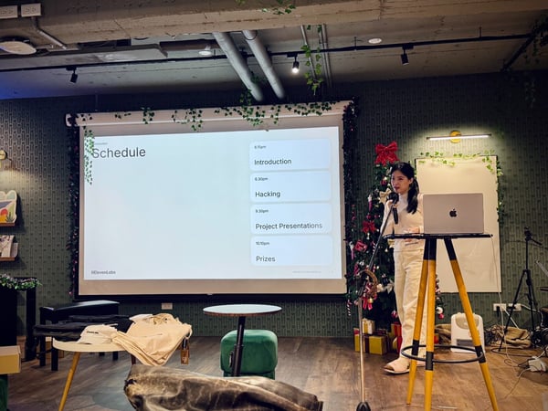 A photograph captures a woman giving a presentation at an indoor event, standing next to a laptop on a tripod table while a large screen displays the event schedule in a room decorated with greenery and Christmas elements.