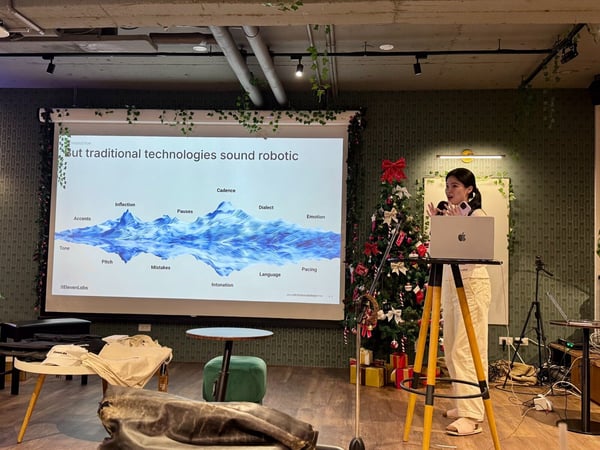 A woman presents in an indoor, festive setting, standing next to a laptop and microphone while a presentation slide about voice technology and its components (like inflection, cadence, and emotion) is projected behind her. The room is decorated with a Christmas tree and ivy vines, suggesting a holiday event or gathering.