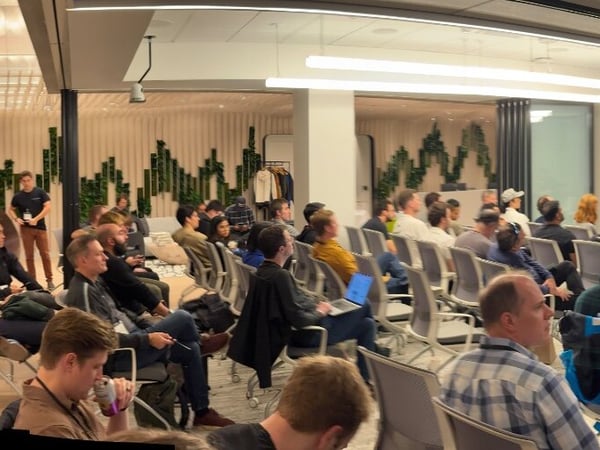 A panoramic photograph captures a large audience seated in a modern conference room attending a technical presentation. A speaker stands at the front addressing the crowd, with multiple large screens visible behind him, one displaying a large blue 'J'.