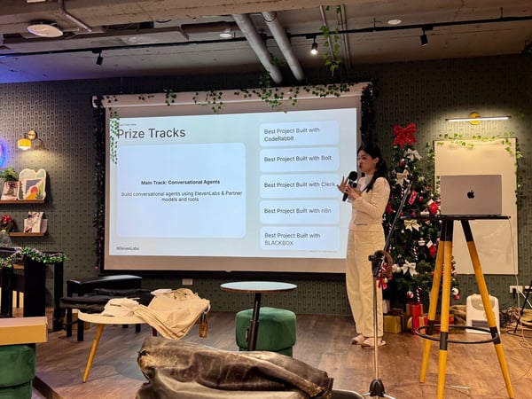 A woman is giving a presentation indoors at an event, standing next to a large screen displaying a slide titled "Prize Tracks" in a room decorated with a Christmas tree and holiday garlands.