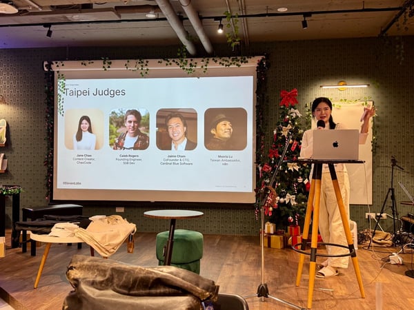 A woman presents indoors at an event, standing beside a laptop and a decorated Christmas tree, with a projected slide listing the "Taipei Judges" and their affiliations visible on a screen behind her.