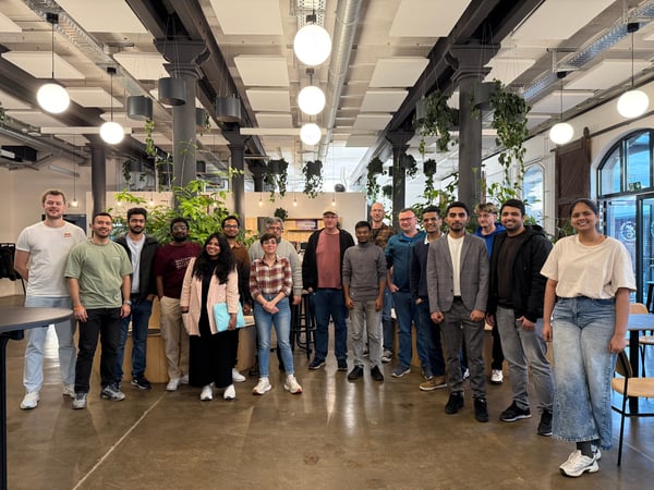 A group of seventeen people posing for a photo in a modern, industrial-style indoor space with high ceilings, hanging plants, and large globe lights.
