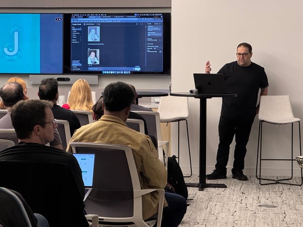 A candid photograph capturing a technical presentation in a modern office setting, showing a male presenter standing next to a laptop and gesturing toward a large dual-screen display while an audience is seated, watching the content.