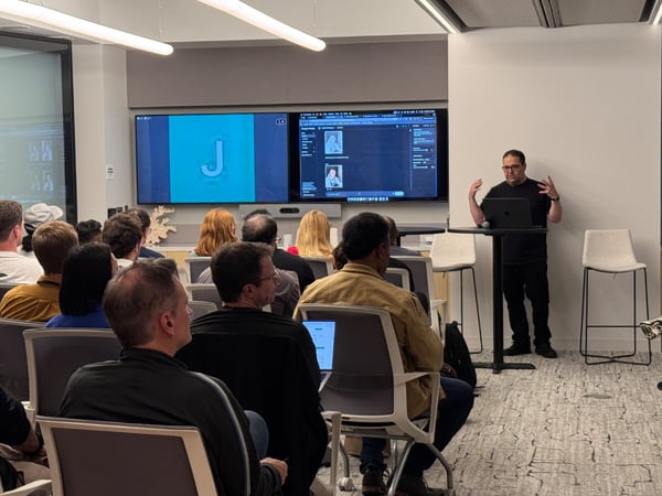 A man is giving a presentation in a modern conference room to a seated audience, with a large screen displaying a capital letter 'J' and technical content behind him.