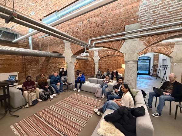 A group of people are gathered in a modern lounge area with vaulted brick ceilings and industrial-style exposed piping. They are seated on grey sofas and chairs, some using laptops, in what appears to be a casual meeting or working session.