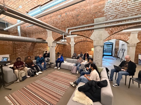 A group of people are gathered in a modern, industrial-style room with exposed brick arches and large overhead pipes. They are sitting on grey sofas and chairs, some using laptops, in what appears to be a casual meeting or social gathering.