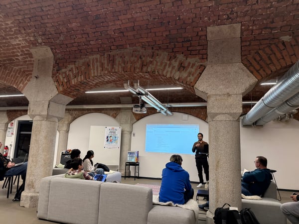 A man stands giving a presentation to a small audience in a unique indoor space featuring a vaulted brick ceiling and large stone pillars.