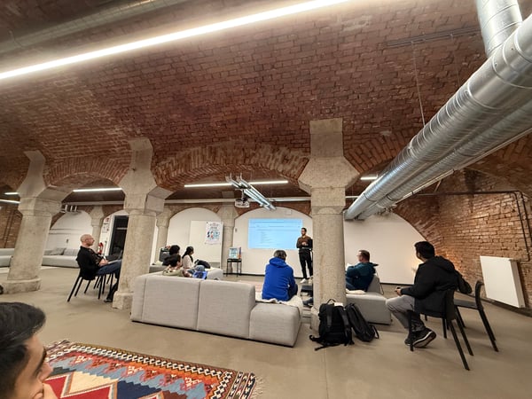 A group of people are gathered in a room with a distinctive brick-vaulted ceiling and stone pillars, attending a presentation or demo being shown on a projector screen.