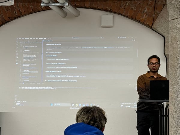 A man is giving a technical presentation, standing next to a laptop with a screen projected onto a white wall showing code and documentation.