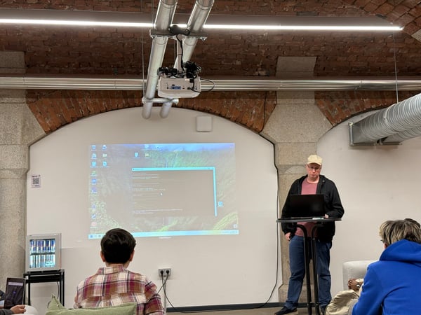 A man stands at a laptop giving a presentation in a room with exposed brick arches, with a projector displaying a computer screen on the wall.