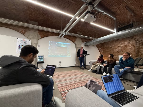 A group of people are gathered in a room with exposed brick walls for a presentation. One person stands at a laptop stand presenting to others who are seated on couches with their own laptops open.