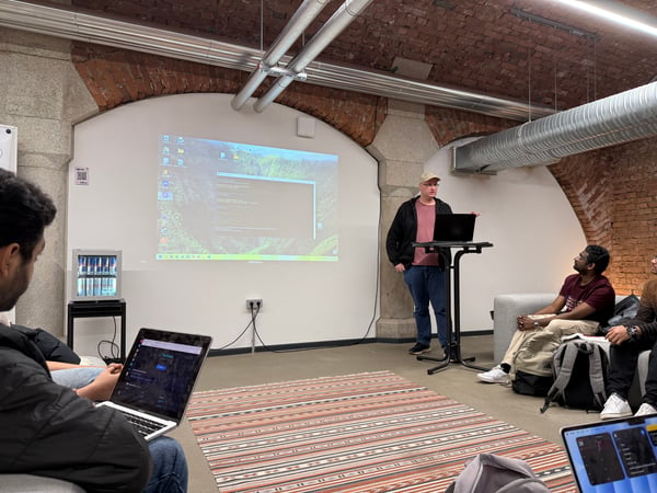 A man stands at a high desk giving a technical presentation or demo to a small group in a room featuring exposed brick arches and industrial piping.