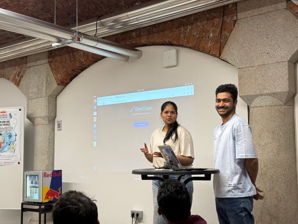 A woman gives a presentation using a laptop on a high table while a man stands beside her, smiling, in an indoor space with exposed brick arches.