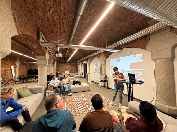 A woman is giving a presentation to a small group of people in a modern office or event space featuring a vaulted brick ceiling.