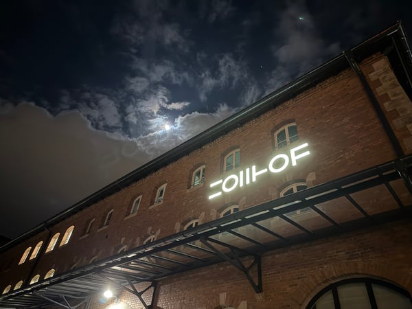 A low-angle night photograph of a red brick building featuring a glowing white 'ZOLLHOF' sign under a cloudy, moonlit sky.
