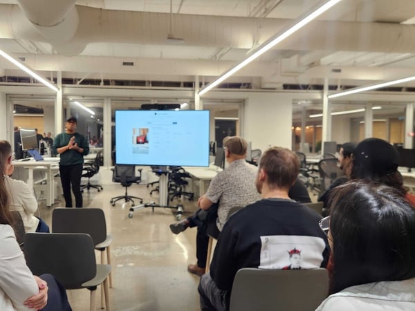 A photograph capturing a presentation or meeting taking place in a modern, open-plan office setting, where a presenter stands before a large screen while an audience is seated facing him.