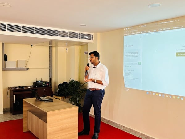 A man wearing a white shirt and dark pants is giving a presentation or software demonstration indoors, standing next to a wooden table while holding a microphone. Behind him, a large projection screen displays a web interface for an 'AI Assistant' application.