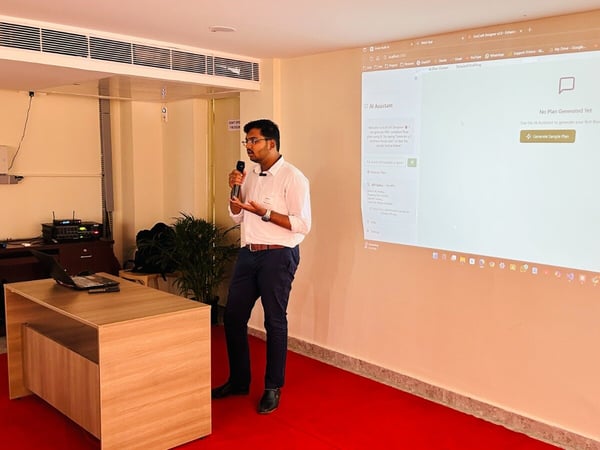 A man wearing a white shirt and dark pants is giving a presentation or technology demonstration, speaking into a microphone while standing beside a large projected screen displaying a web application labeled "AI Assistant."