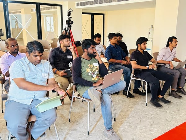 A candid photograph captures a group of men seated in a well-lit indoor conference or training room, attending an event or presentation. Several attendees are focused on laptops or tablets, and a camera setup on a tripod is visible in the background.