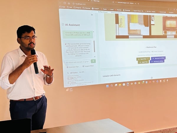 A man wearing glasses and a white shirt is giving a presentation or software demonstration, holding a microphone while gesturing with his free hand. Behind him, a large screen projects a web application interface featuring an AI-assisted architectural design (a 2 Bedroom Plan).
