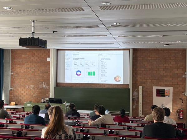 A lecture hall at Eindhoven University of Technology where an audience is watching a presentation of a data dashboard on a large screen.