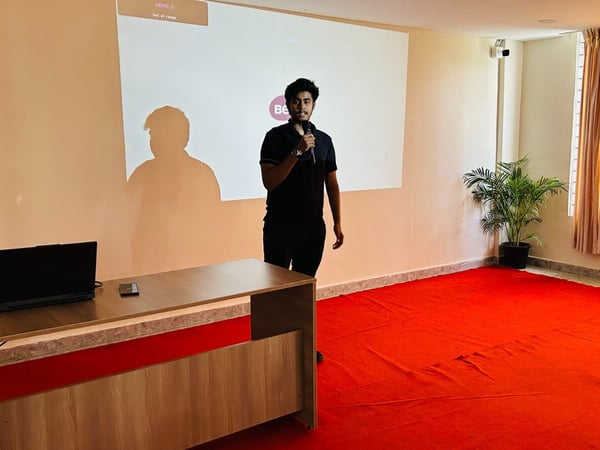 A young man wearing all black stands in a presentation room with a bright red carpet, holding a microphone while facing the audience in front of a large projection screen.