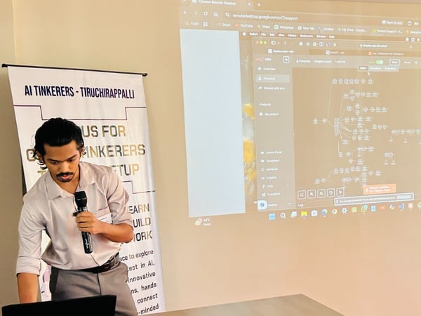 A man is giving a presentation or technical demonstration indoors, standing next to a promotional banner while a complex diagram or interface is projected onto a large screen behind him.
