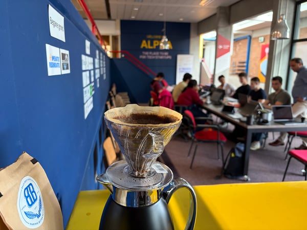 A close-up of a pour-over coffee dripper on a carafe in the foreground, with a blurred background showing people working in a modern coworking space called ALPHA.