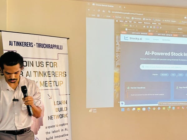 A young man is giving a presentation indoors, holding a microphone, while standing next to a banner for the 'AI Tinkerers - Tiruchirappalli Meetup.' Behind him, a projector screen displays a website interface for 'AI-Powered Stock Insights' (Stocky.ai).