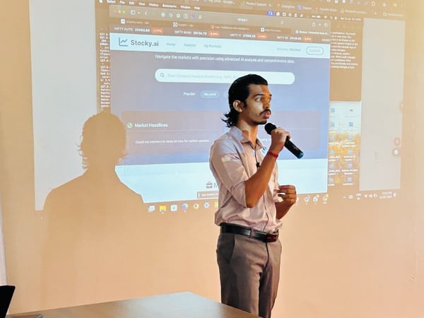 A photograph capturing an indoor event where a man is giving a presentation, holding a microphone, in front of a large projection screen displaying the Stocky.ai website and next to a banner for the 'AI Tinkerers Trichy Meetup'.
