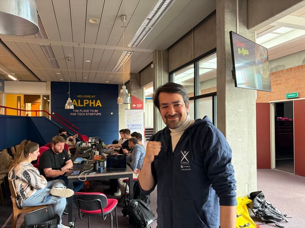 A man in a navy blue hoodie poses with a clenched fist in the foreground of a modern coworking space, where several people are working at a long table with laptops.