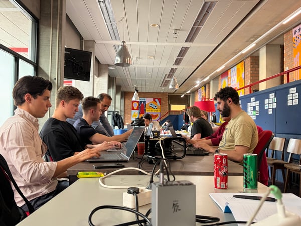 A group of young adults are focused on their laptops while sitting at long tables in a modern, industrial-style workspace.