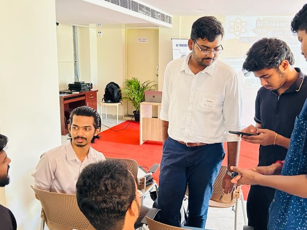 A candid photograph captures a group of young men gathered in an indoor event space, engaged in a discussion, possibly reviewing a small electronic device or phone during a technical workshop.