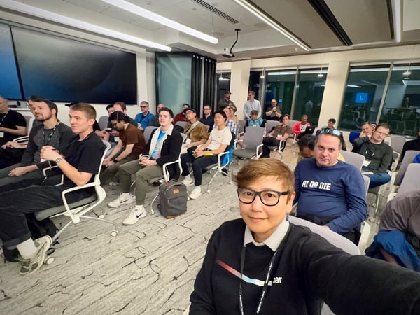 A selfie taken indoors captures a large audience seated in a modern conference room, attending a presentation or meeting, with a large, dark screen visible in the background.