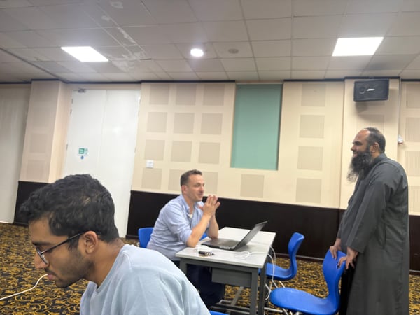 A candid photograph showing three men in an indoor setting, likely a classroom or meeting room, engaged in a discussion around a laptop.