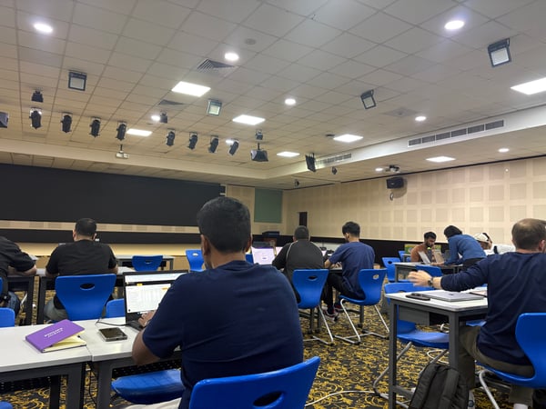 A group of people are seated at desks in a large room, likely a lecture hall or conference space, with many using laptops and facing a large screen at the front.