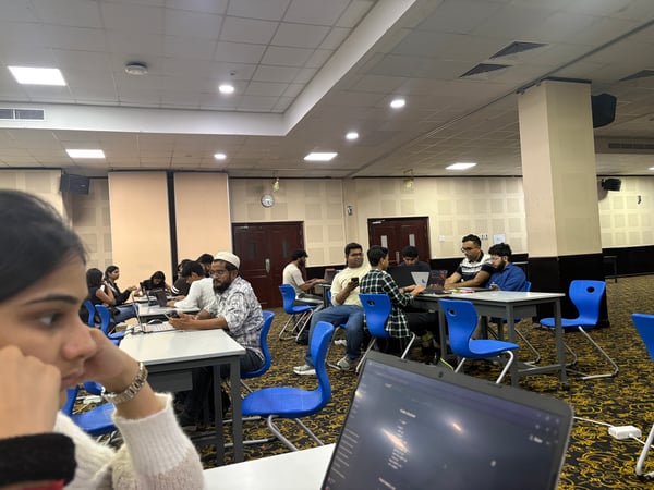 A group of individuals are seated at tables in a large hall, focused on their laptops during what appears to be a workshop or study session.