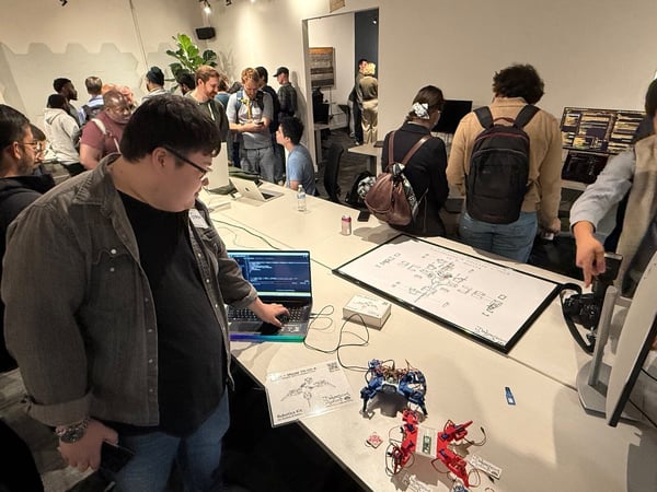 A candid photograph of a tech event or demo day where attendees are gathered around a table featuring a laptop displaying code, a technical diagram, and two small, multi-legged robots.