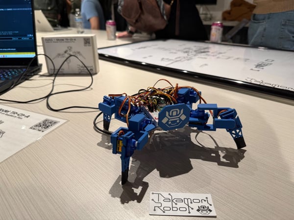 A close-up photograph of a small blue hexapod robot (Model TR-01-A) with exposed wiring, displayed on a light-colored table during what appears to be a technical demonstration or workshop.