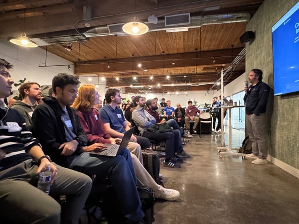 A photograph capturing a presentation event indoors, showing a speaker standing on the right addressing a large, seated audience in a venue with an industrial aesthetic, featuring exposed ceilings and concrete floors. Several audience members in the foreground are attentively watching the speaker and the large blue screen displaying text.