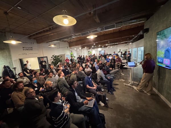 A photograph capturing a large audience attending an indoor presentation or talk, where a woman is speaking into a microphone near a large screen displaying video content.