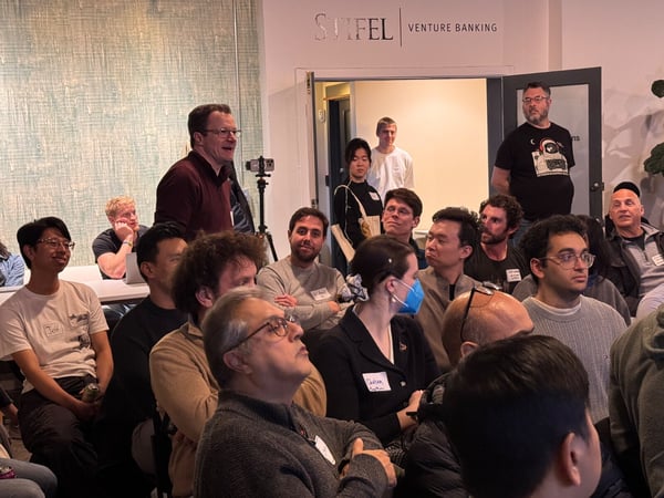 A candid photograph showing a large group of people gathered indoors for a presentation or meeting, with one man standing and speaking near the center of the frame while the audience listens. The wall above a doorway features corporate branding reading "STIFEL | VENTURE BANKING".