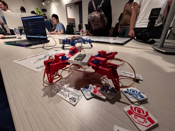 A photograph capturing an indoor technical event or workshop, showcasing red and blue 3D-printed robotic prototypes and branded keychains displayed on a light-colored table, with attendees working on laptops and observing in the background.