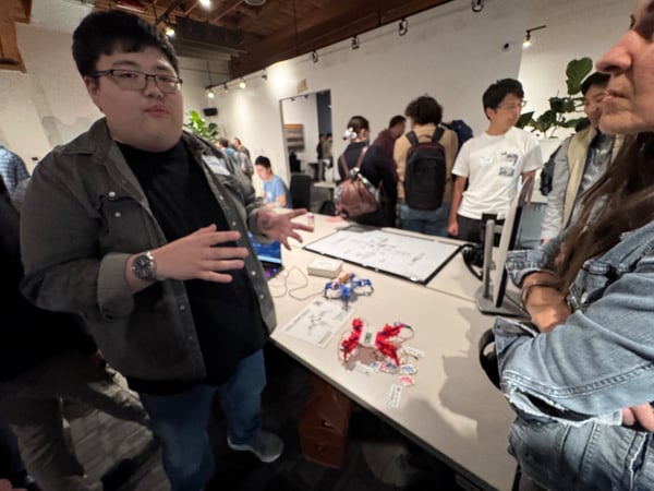 A candid photograph taken indoors at an event, showing a young man gesturing while presenting a project involving small electronic components displayed on a table to a group of attendees.