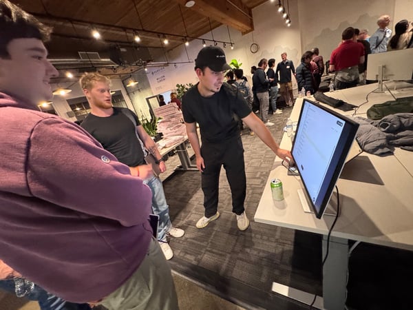 A candid photo taken indoors at a professional or networking event, showing three young men gathered around a large monitor displaying data while a larger group socializes in the background.