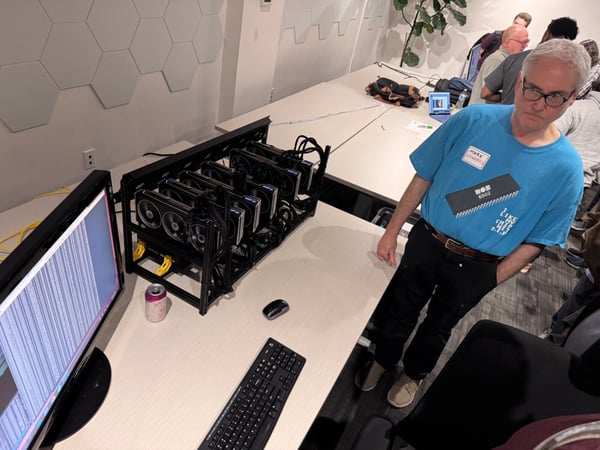 A candid indoor photograph capturing a technical meeting or workshop, featuring a man standing next to a multi-GPU computing rig setup on a desk, with other attendees visible in the background.