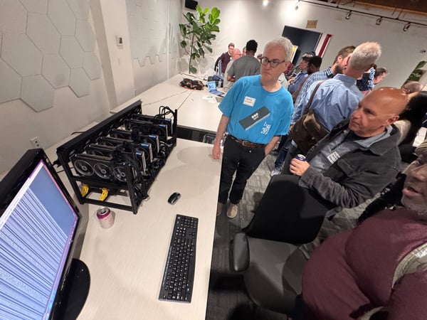 A candid photograph taken indoors at a tech event or meetup, showing a group of people gathered around a table where a multi-GPU mining rig setup is being demonstrated, connected to a monitor displaying data.