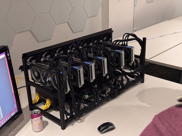 A photograph showing a multi-GPU computing rig, likely used for cryptocurrency mining, set up on a white desk in an indoor office environment. A can of Waterloo sparkling water and a computer mouse are visible next to the complex hardware setup.