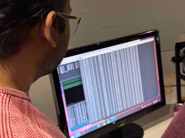 A close-up, over-the-shoulder photograph shows a person wearing a pink knitted sweater and glasses, intently focused on a computer monitor displaying dense columns of data or code.